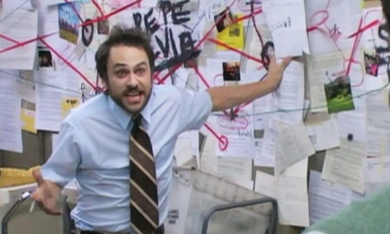 A still from It's Always Sunny in Philadelphia showing Charlie Day standing in front of a cork board covered in clues and red yarn. He's holding a cigarette and wearing a blue short sleeved button up shirt. His hair is disheveled and he appears very annoyed.