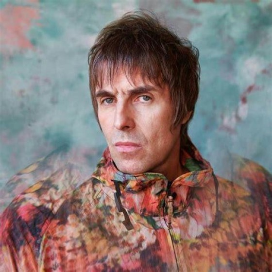 A headshot of Liam Gallagher from the band Oasis. He's wearing a tie-dye style printed windbreaker and posing like he's at Sears in 1985. His hair is from 1964.