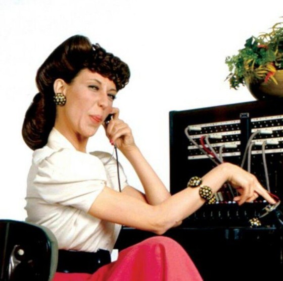 Ernestine, a character portrayed by Lily Tomlin. A phone operator in 40s-50s American clothing sitting at a switchboard with a headset on. Her tongue is sticking out.