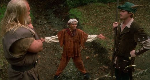 A scene from Robin Hood: Men In Tights showing Ahchoo (Dave Chappelle) straddling a small stream, framed slightly below a small bridge and by Robin of Loxley (Cary Elwes) and Little John (Eric Allan Kramer). 