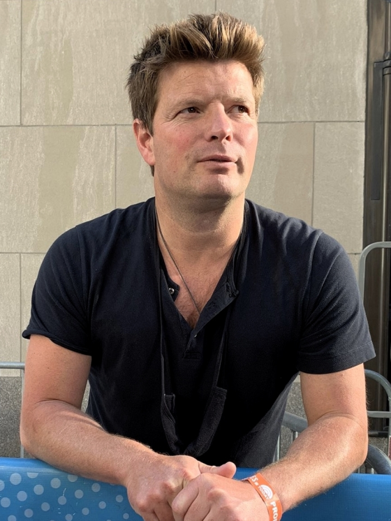 Coldplay's Phil Harvey wearing a black t-shirt and orange wrist band, looking off camera.